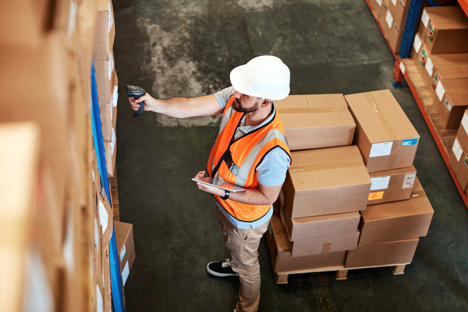 A warehouse worker, equipped with a white hard hat and orange safety vest, scans boxes on a shelf using a handheld scanner while holding a tablet displaying UPS shipping details. Nearby, cardboard boxes are stacked on pallets, ready for dispatch as the warehouse floor bustles with activity.