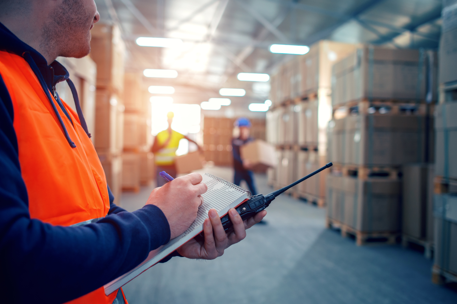 A person in an orange safety vest holds a notepad and a walkie-talkie in a bustling warehouse, coordinating an efficient UPS pickup. In the background, two helmeted workers handle boxes between tall stacks of pallets, all under bright overhead lights.