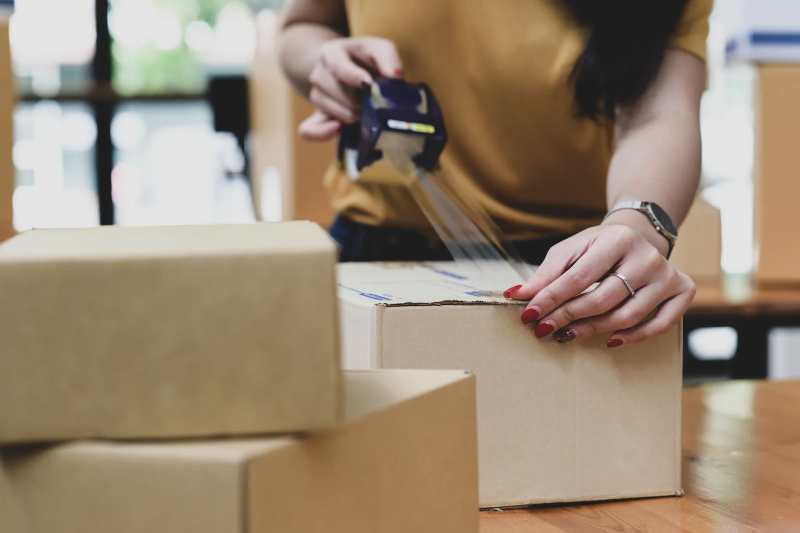 A person with long dark hair seals a cardboard box with packing tape on a table, surrounded by other boxes. The scene, set in a bright indoor area, hints at e-commerce logistics hustle during UPS Peak Season, readying shipments amid soaring demand.